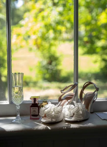 Bridal shoes with pearl wedding heels on a window sill, styled beside a perfume bottle and champagne flute in soft window light