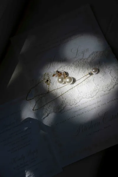 Bridal jewelry flatlay with wedding invitation flatlay, pearl drop earrings and a gold pearl necklace on lace fabric beside stationery in soft light