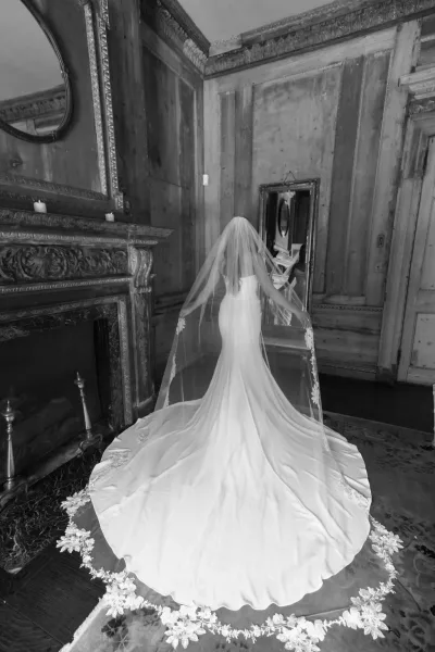 Bridal veil portrait of a bride in a cathedral veil with lace trim and long train, standing in a wood-paneled room by an ornate fireplace and mirror