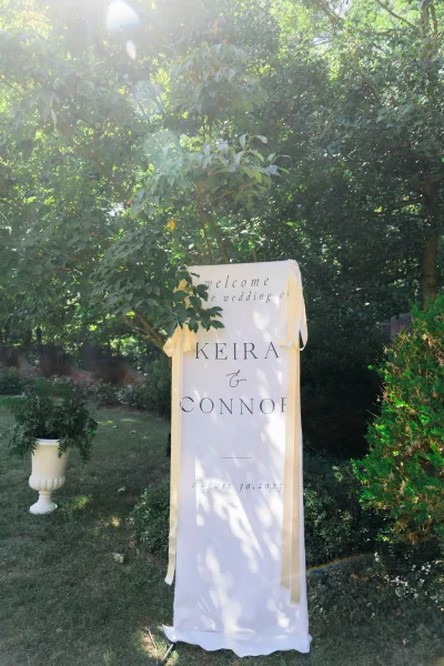 Wedding welcome sign with calligraphy text on draped fabric, satin ribbons on a stand beside an urn planter in a sunlit garden backdrop