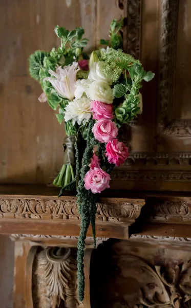 Bridal bouquet of pink and white bouquet blooms with greenery and trailing amaranthus, ribbon-wrapped, resting on a carved wooden mantle