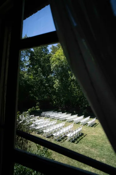 Ceremony seating with white folding chairs arranged in rows on a grassy lawn, framed by a window with curtains and trees beyond