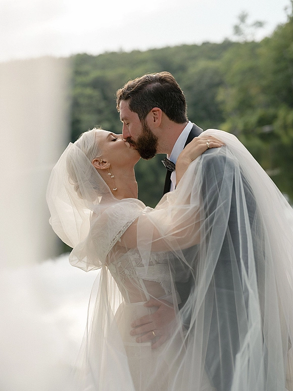 Wedding kiss portrait of bride and groom under a sheer veil, her hand on his shoulder, outdoors by greenery and water