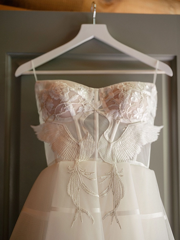 Wedding dress on a hanger with strapless lace corset bodice and embroidered floral appliqué, reflected in a framed mirror under warm wood ceiling
