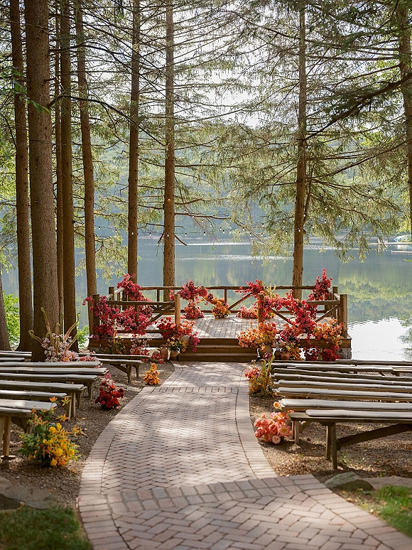 Ceremony setup for an outdoor wedding ceremony with a wooden platform, bright florals, benches, and string lights by a lakeside forest shore