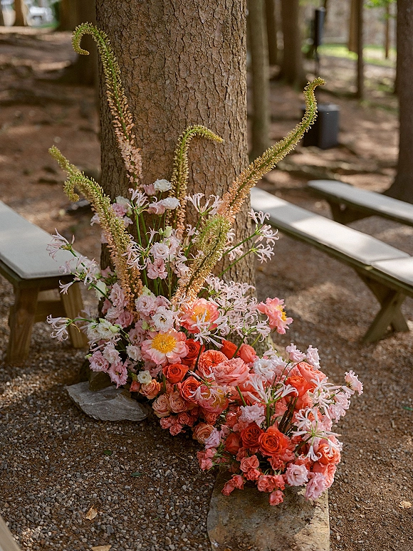 Ceremony aisle florals with coral and pink roses in a grounded arrangement at a tree base beside gravel and wooden benches in forest setting