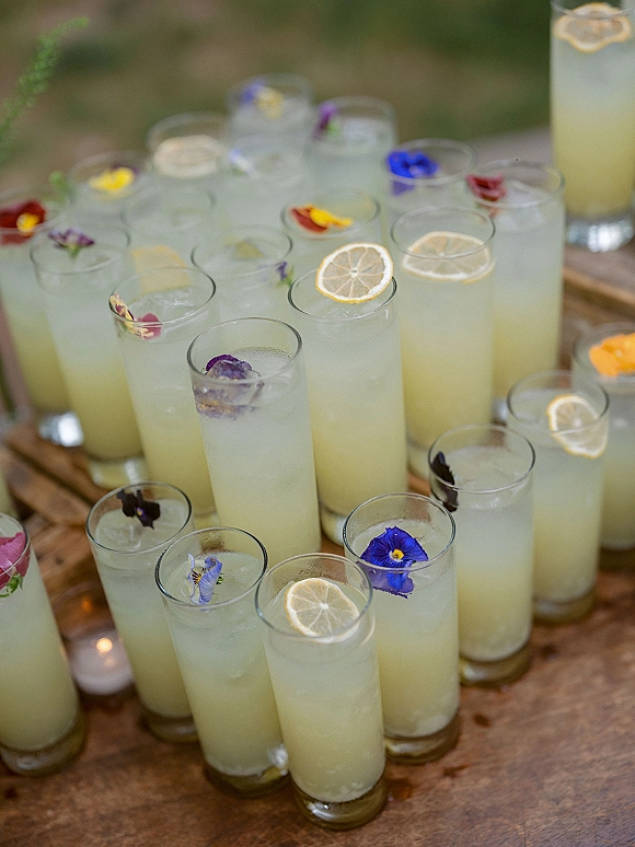 Wedding welcome drinks in tall lemonade glasses with lemon slices and edible flower accents on a wooden tray set on a grassy lawn