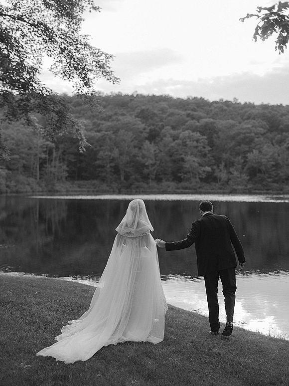 Couple portrait of bride and groom walking away holding hands, veil and dress train flowing by a tranquil lakeshore and trees.