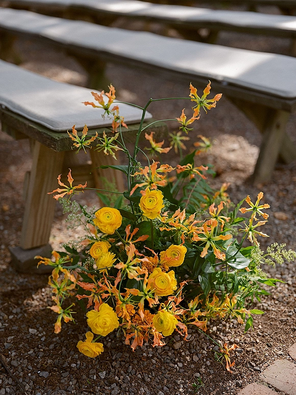 Ceremony aisle flowers with yellow ranunculus and orange gloriosa lilies, lush greenery on gravel beside sunlit wooden benches outdoors