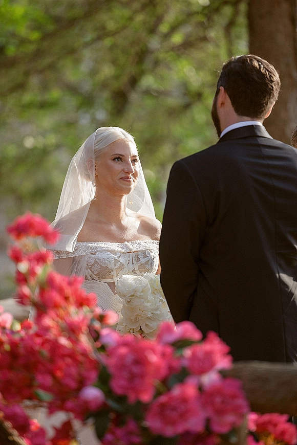 Ceremony moment as bride and groom at altar exchange vows, bride in veil and strapless lace dress holding bouquet amid pink flowers