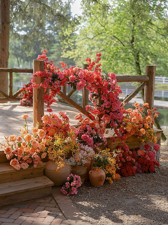 Wedding floral decor with pink and coral blooms in terracotta pots lining wooden steps, with greenery and a sunlit pond behind