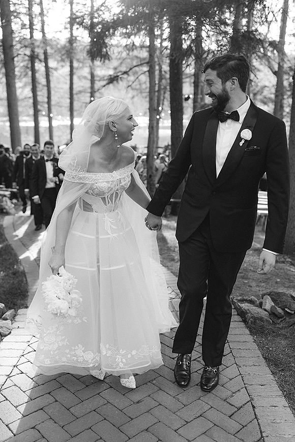 Wedding recessional as bride and groom walk the aisle hand in hand, her veil and white bouquet flowing on a brick path under string lights