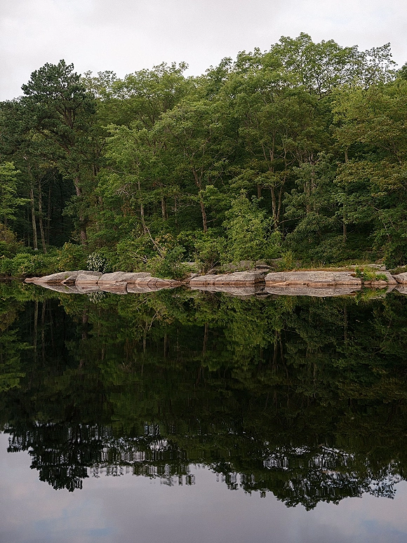 Lake landscape with forest lake reflection, calm water mirroring dense trees and rocky shoreline beneath an overcast sky