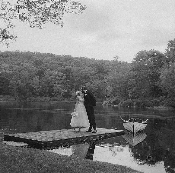 Wedding kiss portrait of bride and groom kissing on a dock, her bouquet in hand, a rowboat beside them on a calm lake backdrop