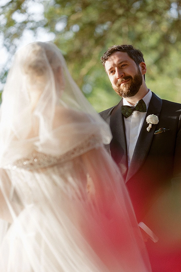 First look moment as bride approaches from behind her veil, groom in tuxedo turning with an emotional reaction in sunlit trees