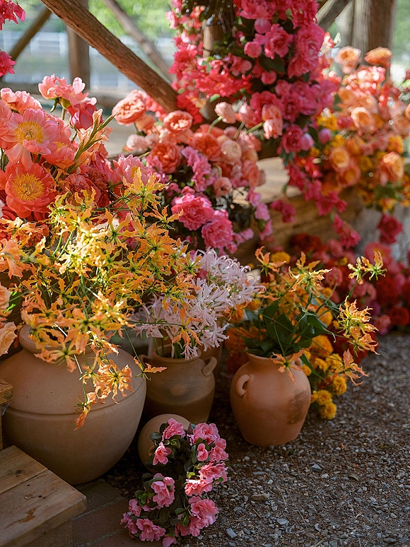 Wedding floral arrangement in terracotta vases with bright pink, coral, orange, and yellow blooms with greenery on gravel outdoors