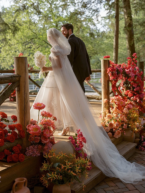 Wedding couple portrait from behind, bride holding bouquet as a long veil trails along a flower-lined wooden deck in natural light