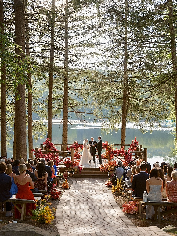 Wedding ceremony with bride and groom at a floral arch, bride in veil and groom in suit on a wooden deck by a lake and mountains