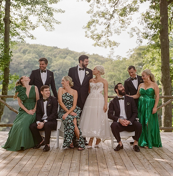Wedding party portrait of bride with bridesmaids and groom with groomsmen seated on benches on a wood deck by a lake and trees, laughing