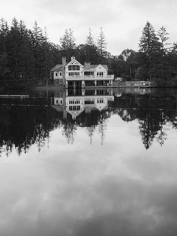 Wedding venue exterior of a lakeside wedding venue with a balcony, reflected in calm lake water beneath evergreen trees and cloudy sky