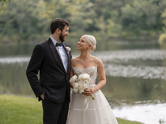 Couple portrait of bride in strapless lace dress holding a white bouquet with groom in black tuxedo, standing by a lakeside forest shoreline