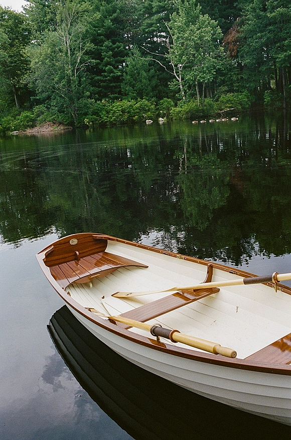 Rowboat photo of a classic wooden boat with oars drifting on a calm lake, with forest trees and rocky shoreline reflected in water