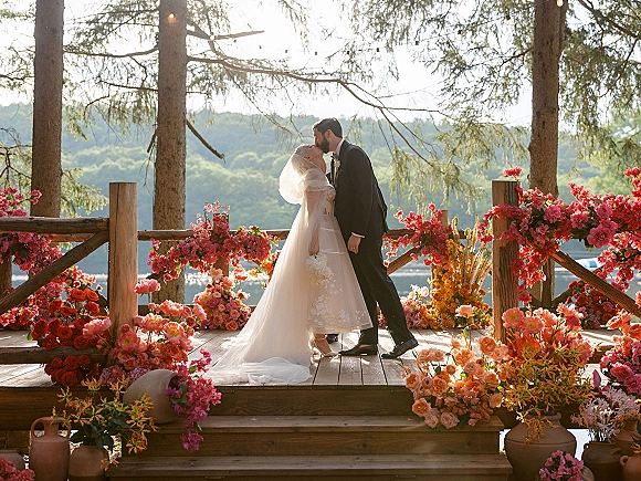 Wedding kiss as bride in veil and groom in tuxedo embrace on a floral deck with string lights, pine trees, lake and mountains behind