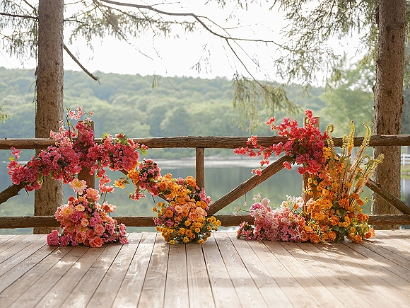 Wedding ceremony backdrop with colorful ceremony flowers on a wooden deck railing, set against a lakeside view of forested hills