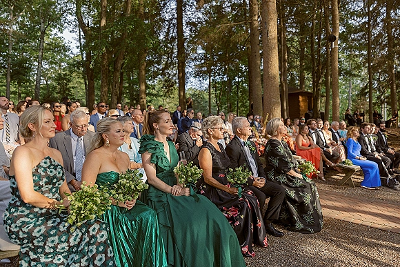 Wedding guests seated on wooden benches, outdoor wedding guests in emerald green dresses and suits holding bouquets in a sunlit forest setting