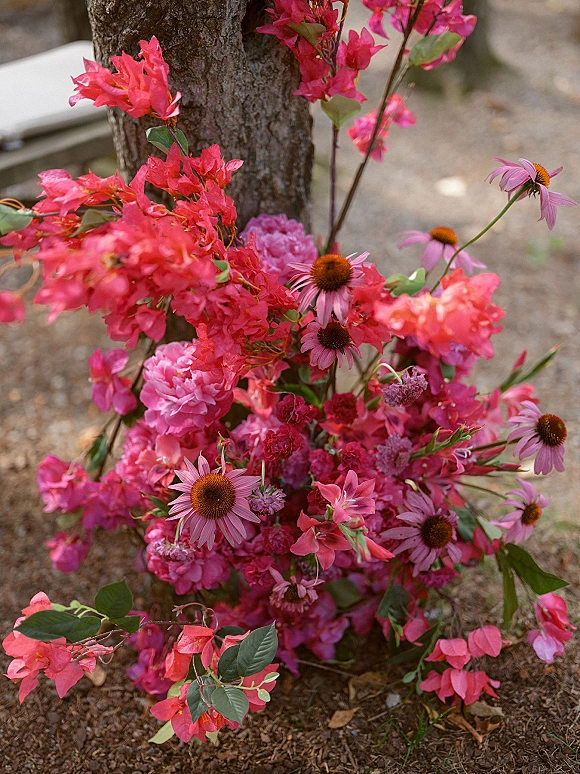 Wedding floral installation with pink florals and bougainvillea branches arranged at a tree base on dirt in an outdoor garden setting