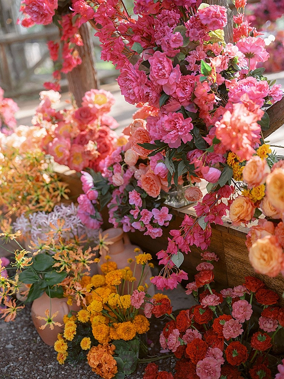 Ceremony florals in a wedding floral installation with pink, coral, orange, and yellow blooms and greenery in terracotta and glass vases outdoors
