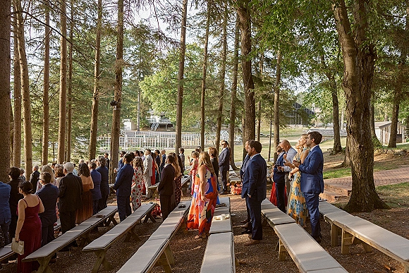 Wedding guests in suits and colorful dresses stand beside bench seating during a forest ceremony in an outdoor clearing with trees and gravel ground