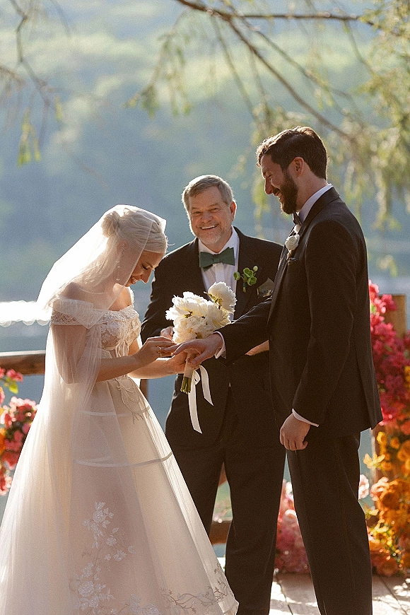 Wedding vows as bride and groom exchange rings on a lakeside deck, bride in lace dress and veil holding bouquet, mountains behind