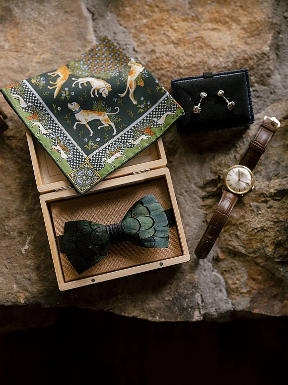 Groom accessories flat lay with green bow tie in a wooden box, patterned pocket square, cufflinks, and wristwatch on stone surface