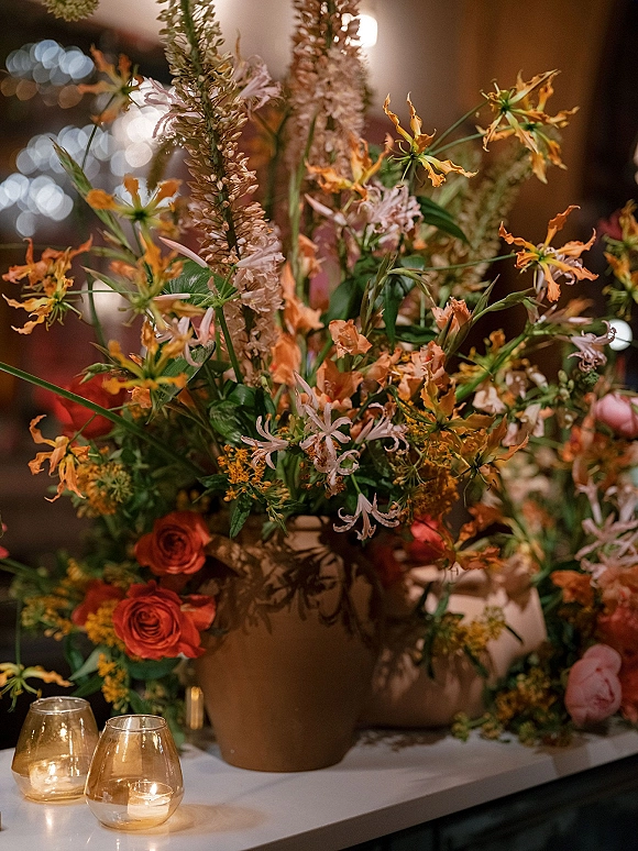 Wedding centerpiece with wildflower wedding centerpiece in a ceramic vase, roses and greenery beside votive candles in warm bokeh-lit reception room