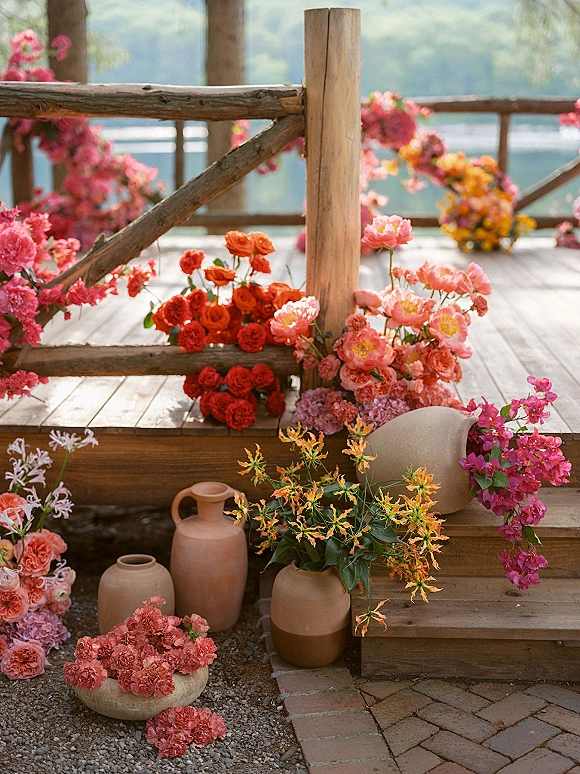 Wedding ceremony florals and ceremony aisle florals in terracotta vases with coral and pink blooms on a wooden deck beside a lake backdrop