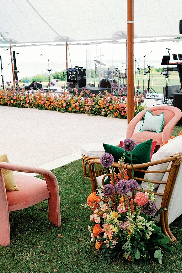 Reception lounge area with wedding lounge seating, rattan sofa and floral arrangements under a white tent canopy beside band stage equipment
