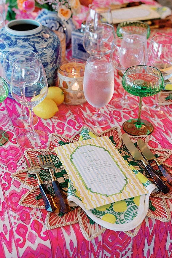 Reception tablescape with colorful wedding table decor on a pink patterned tablecloth, lemons, blue-and-white vase florals, candles, and green coupe glasses