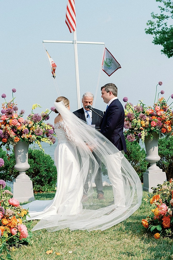 Wedding ceremony with bride and groom exchanging vows under a floral arch, bride’s long veil flowing on a coastal lawn by the ocean