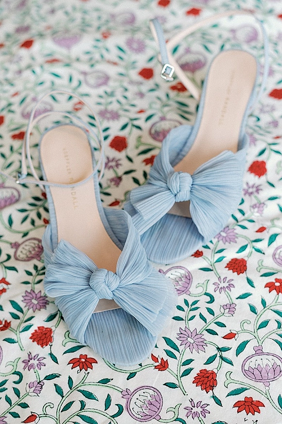 Bridal shoes in a flat lay, blue bridal heels with pleated bow and ankle straps resting on floral patterned fabric backdrop