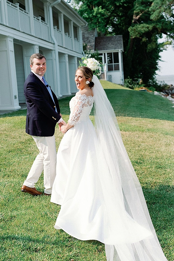 Couple portrait of bride and groom holding hands, bride looking back with long veil and bouquet on a waterfront lawn near a white house