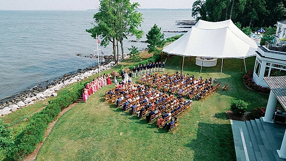 Outdoor wedding ceremony under a white sailcloth tent with floral aisle and wooden chairs on a rocky oceanfront lawn with bridal party lined up