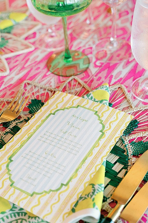 Wedding place setting with a wedding menu card on a patterned napkin, rattan placemat, gold flatware, and green goblet on pink tablecloth