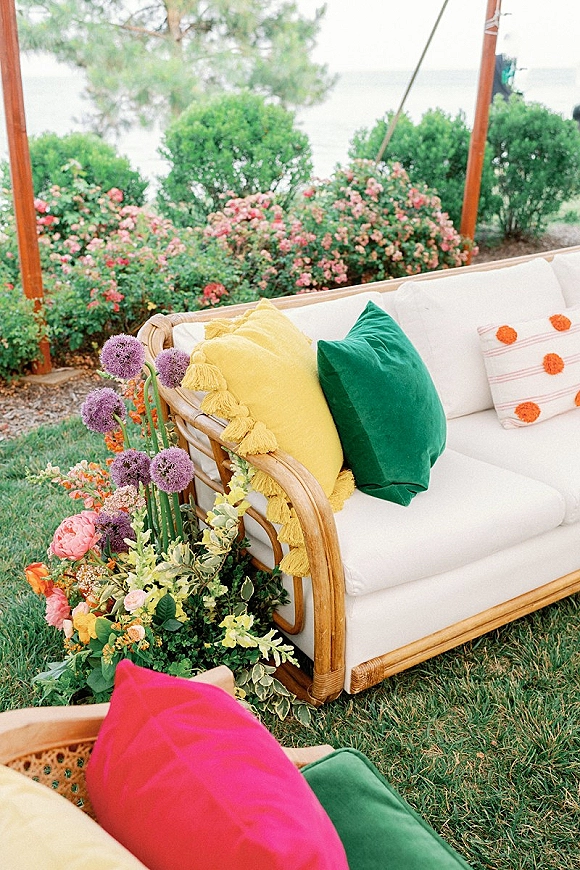 Wedding lounge seating with a rattan wedding sofa, white cushions, colorful throw pillows, and a floral arrangement on a garden lawn