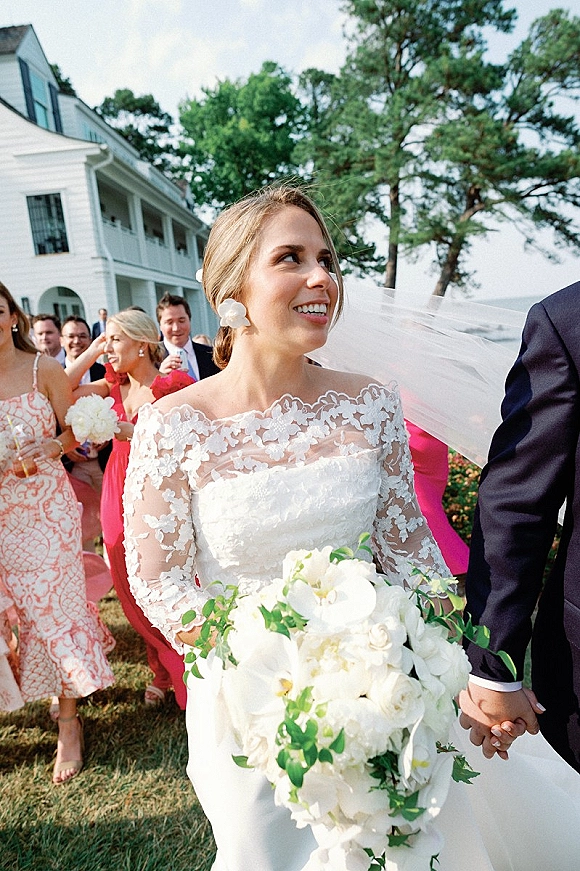 Wedding recessional as bride and groom walk away holding hands, her veil blowing, holding white orchid bouquet on a sunny lawn at a white house venue
