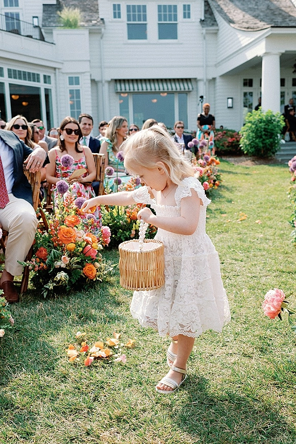 Flower girl tossing petals from a wicker basket down an outdoor aisle lined with roses, with guests on wooden chairs by a white house