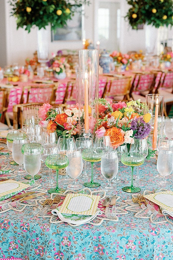 Reception tablescape with colorful wedding tablescape details, floral centerpiece, tall cylinder candles, gold flatware, and green coupe glasses indoors
