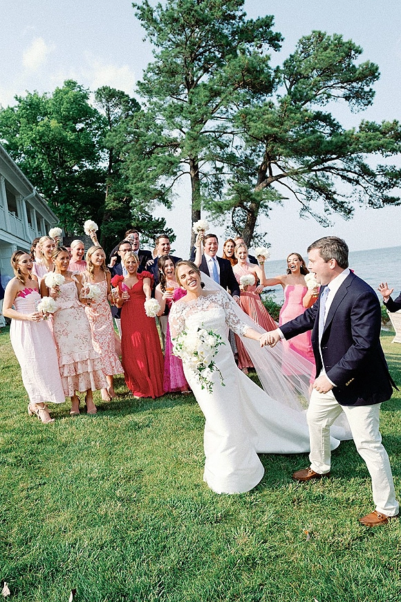 Wedding party photo of bride and groom with wedding party on a lawn by the shoreline, bride in veil holding a white rose bouquet