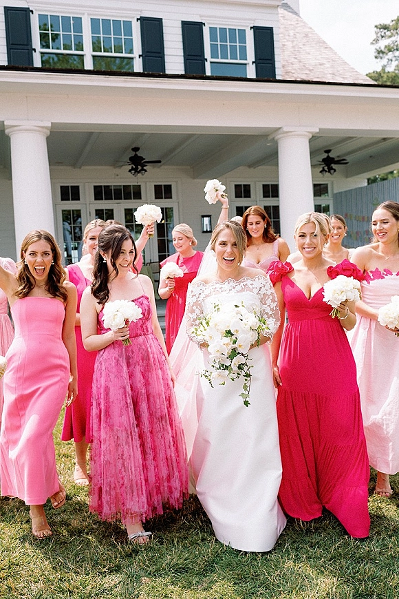 Bride with bridesmaids walking in pink dresses, holding bouquets beside a white house with porch columns, bride in veil and white gown