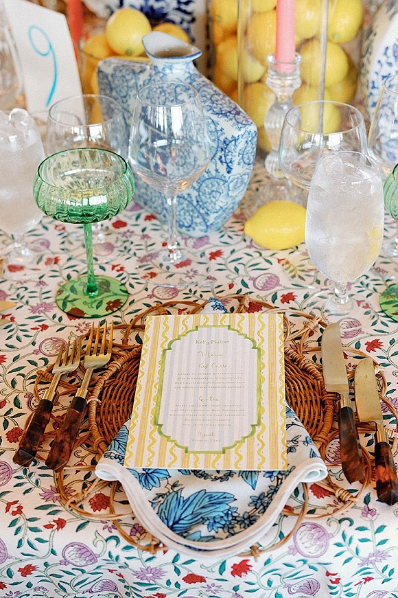 Reception tablescape with a wedding place setting on patterned cloth, rattan charger, printed menu, pink taper candle, and lemons in a jar
